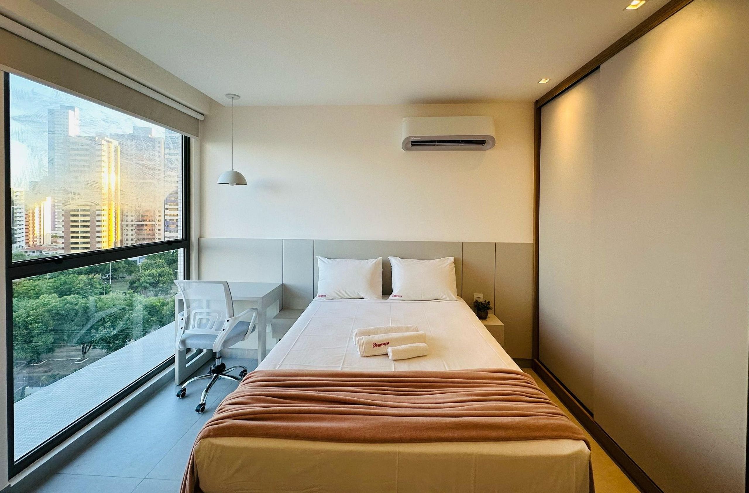A hotel room with a large window, a bed with white linens, a desk, and a chair.