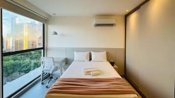 Quarto de hotel com uma janela grande, cama com roupa de cama branca, escrivaninha e uma cadeira.