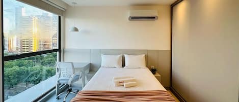 A hotel room with a large window, a bed with white linens, a desk, and a chair.