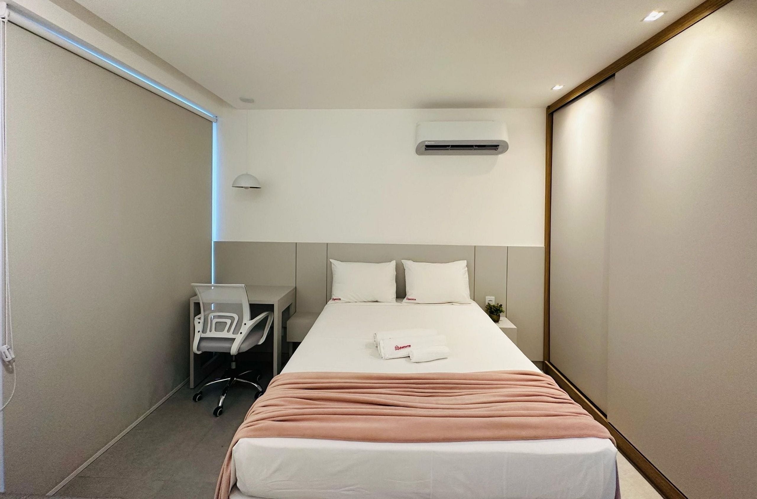 A hotel room with a bed, bedside table, chair, and an air conditioning unit.