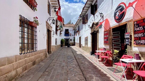 A narrow cobblestone street with white buildings, red chairs, and tables outside a sushi restaurant. - Amira's House Cusco (Cusco)