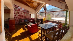 A cozy interior with a wooden ceiling, a brick wall adorned with pictures, a seating area with red cushions, and a view of a cityscape through large windows. - Amira's House Cusco (Cusco)