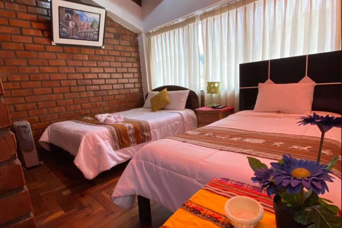 Ein Hotelzimmer mit zwei Betten, einer Ziegelwand, einem Fenster mit Vorhängen und einer Blumenvase.