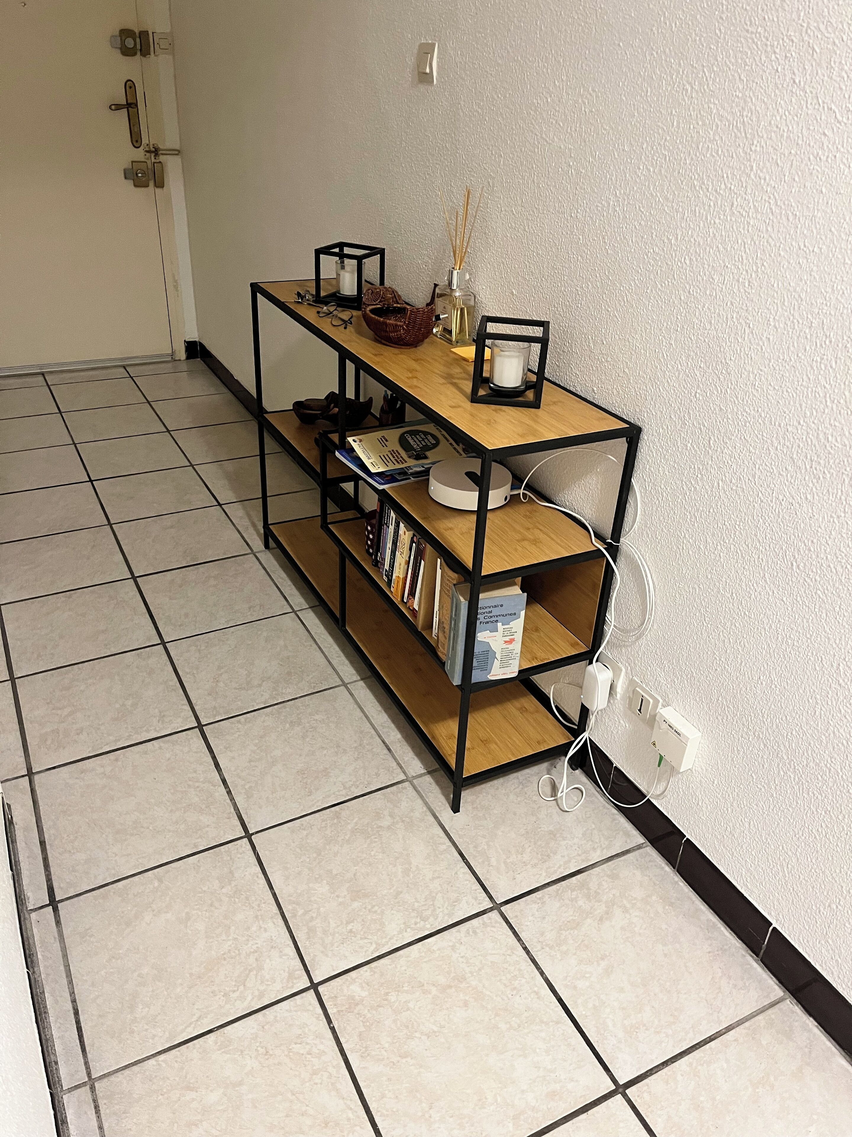 A room with a tiled floor, a wooden bookshelf with books and decorative items, and a wall socket.