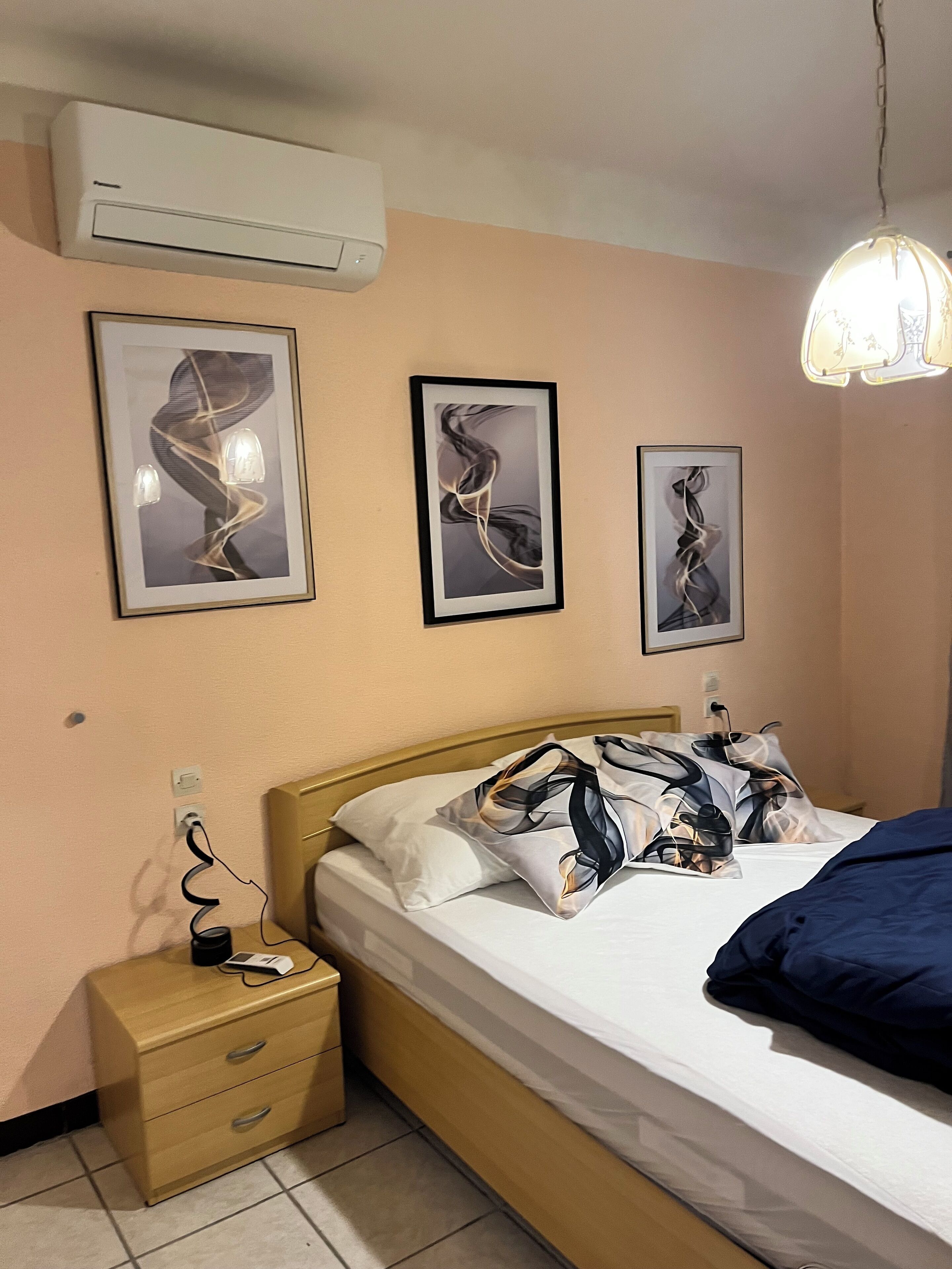 A bedroom with a bed, bedside table, wall-mounted air conditioner, and framed artwork.