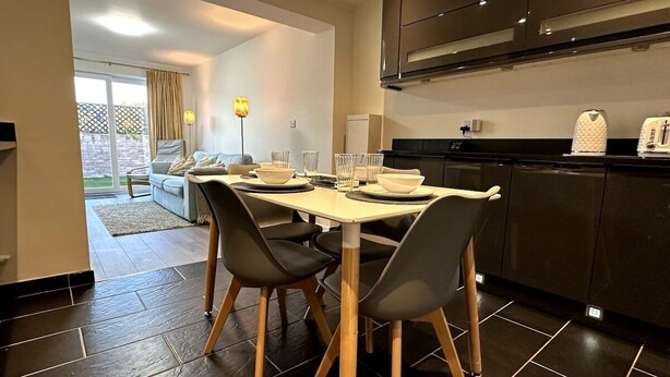 A modern kitchen with dark tiles, a dining table with chairs, and a view to a living area with a sofa and a lamp.