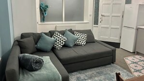A modern living room with a grey sofa, patterned cushions, a small table, and a refrigerator.