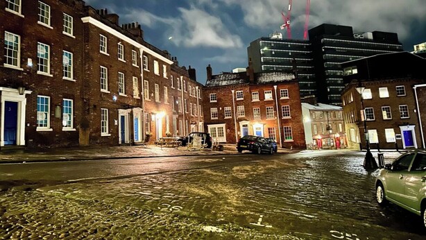 A cobblestone street with historic buildings, a modern skyscraper, and cranes in the background.