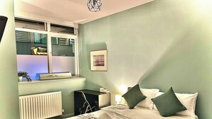 A bedroom with a bed, a desk, a radiator, a window with blinds, and a picture on the wall.