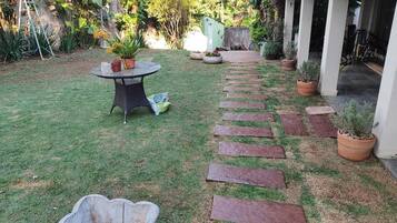 Un jardín con un camino empedrado, una mesa redonda con sillas y plantas en macetas.