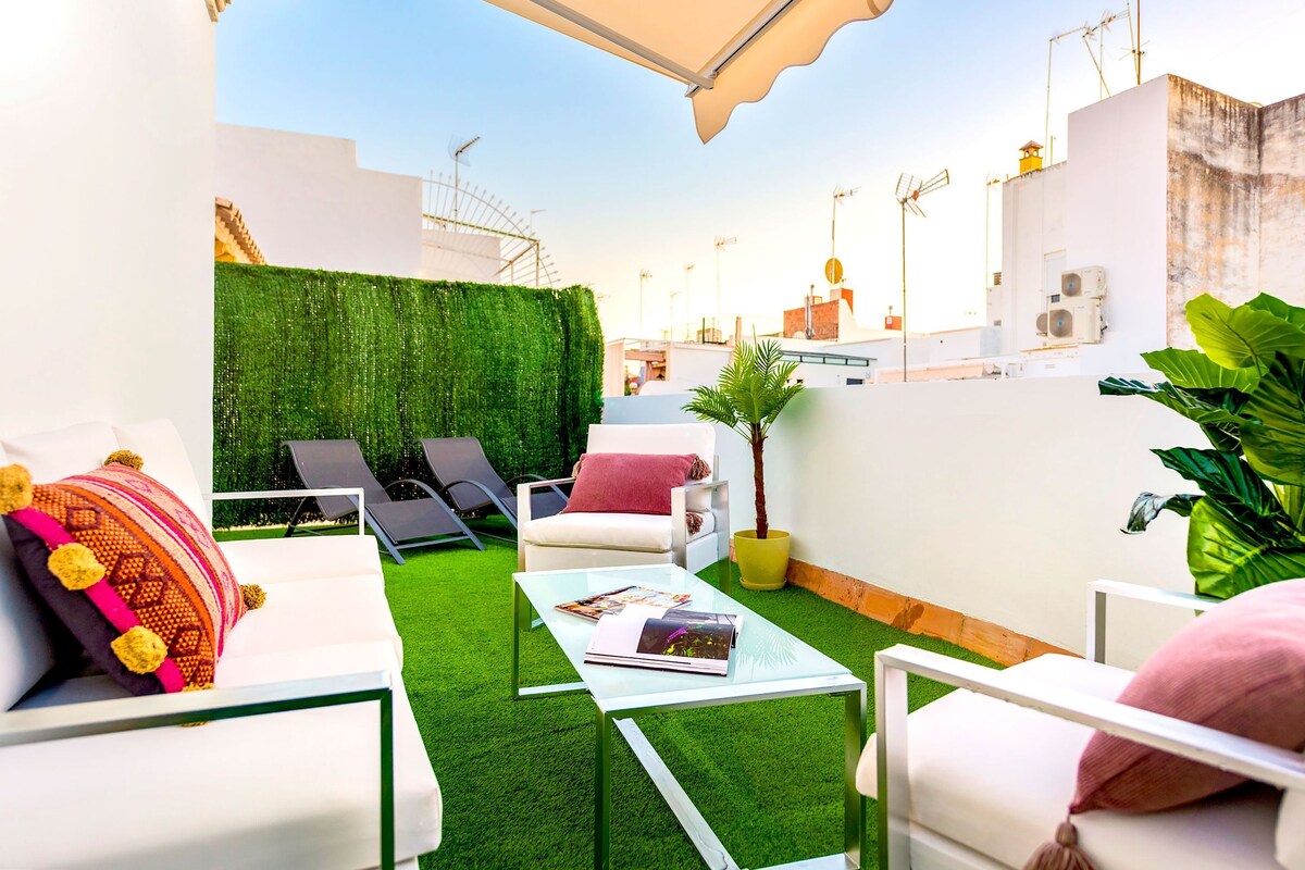 Eine Dachterrasse mit weißen Möbeln, künstlichem Rasen in Grün und einer weißen Wand mit Antennen.