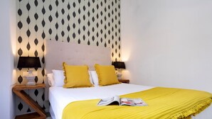 A neatly made bed with a yellow blanket, two pillows, and a magazine on it. There are two bedside lamps and a nightstand with a lamp. The wall has a geometric pattern. - Private Terrace 5 min Walk to Cathedral 2Bd Apartment Galera II (Seville)