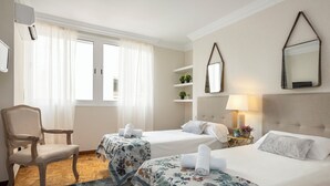 Habitación de hotel con dos camas, una silla, una mesita con lámpara y una ventana con cortinas.