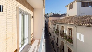 Une rue étroite en milieu urbain, bordée de bâtiments des deux côtés, avec des balcons ornés de plantes, un ciel dégagé et un trottoir pour les piétons.