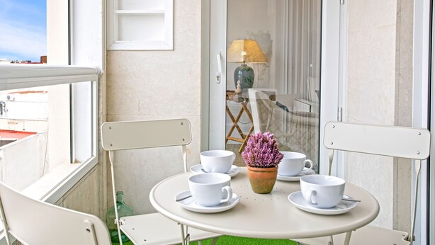 Une petite table ronde, entourée de chaises blanches, et sur laquelle sont posées quatre tasses et soucoupes blanches. Au centre, on aperçoit un pot de fleurs orné de fleurs violettes.