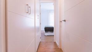 Un couloir étroit aux murs blancs et au plancher en bois mène à une pièce où se trouvent un lit et une fenêtre.