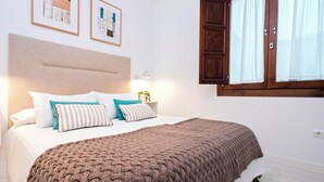 Un dormitorio con una cama, almohadas, una manta de punto, una mesita de noche, una lámpara y una ventana con cortinas.