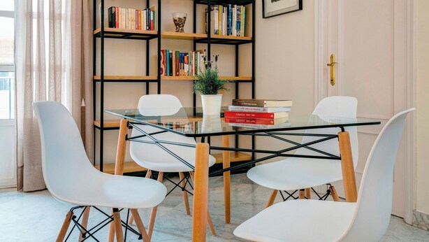 A dining area with a glass table, white chairs, a bookshelf with books and decorative items, and a door with a brass handle. - Spacious Bright With Excellent Location 1 Bd Aptm Tomas de Ibarra (Seville)