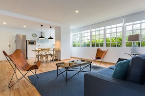 Uma sala de estar moderna com um sofá, cadeiras, uma mesa de centro e vista para uma cozinha e jardim através de grandes janelas.