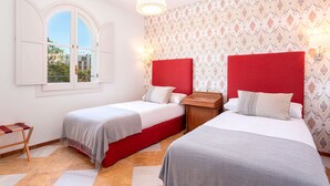 Una habitació amb dos llits, una finestra amb persianes, paper pintat amb dibuixos i una làmpada penjant.