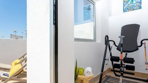 Ein Zimmer mit Fenster, einem Sessel, einem kleinen Tisch mit Handtuch und Hanteln sowie einer Pflanze.