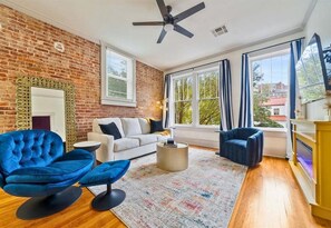 A living room with a brick wall, a ceiling fan, a large window with curtains, a fireplace, and a sofa.