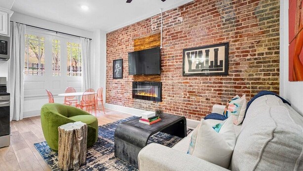 A living room with a brick wall, a fireplace, a green chair, a gray sofa, and a dining area with a white table and red chairs. - Stylish Loft Retreat in Downtown Hot Springs (Hot Springs)
