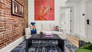 A living room with a brick wall, a white sofa, a black coffee table, and a colorful abstract painting. - Stylish Loft Retreat in Downtown Hot Springs (Hot Springs)