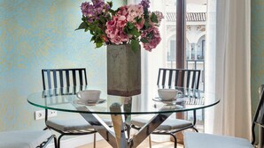 Un coin repas avec une table en verre, deux chaises, un vase de fleurs et une vue sur un bâtiment par la fenêtre.
