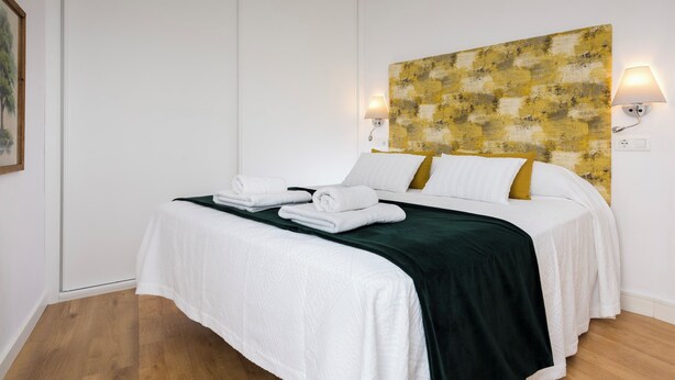 Une chambre d’hôtel avec un lit, du linge de lit blanc, un plaid vert et un tableau abstrait jaune et blanc accroché au mur.