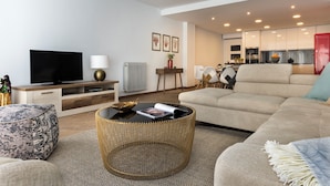 Ein modernes Wohnzimmer mit Sofa, Couchtisch und Fernseher. Im Hintergrund ist eine Küche zu sehen.