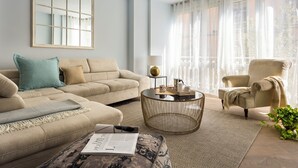 Ein Wohnzimmer mit einem beigefarbenen Sofa, einem runden Couchtisch und einem Sessel. Ein großes Fenster mit transparenten Vorhängen bietet Blick auf viel Grün.