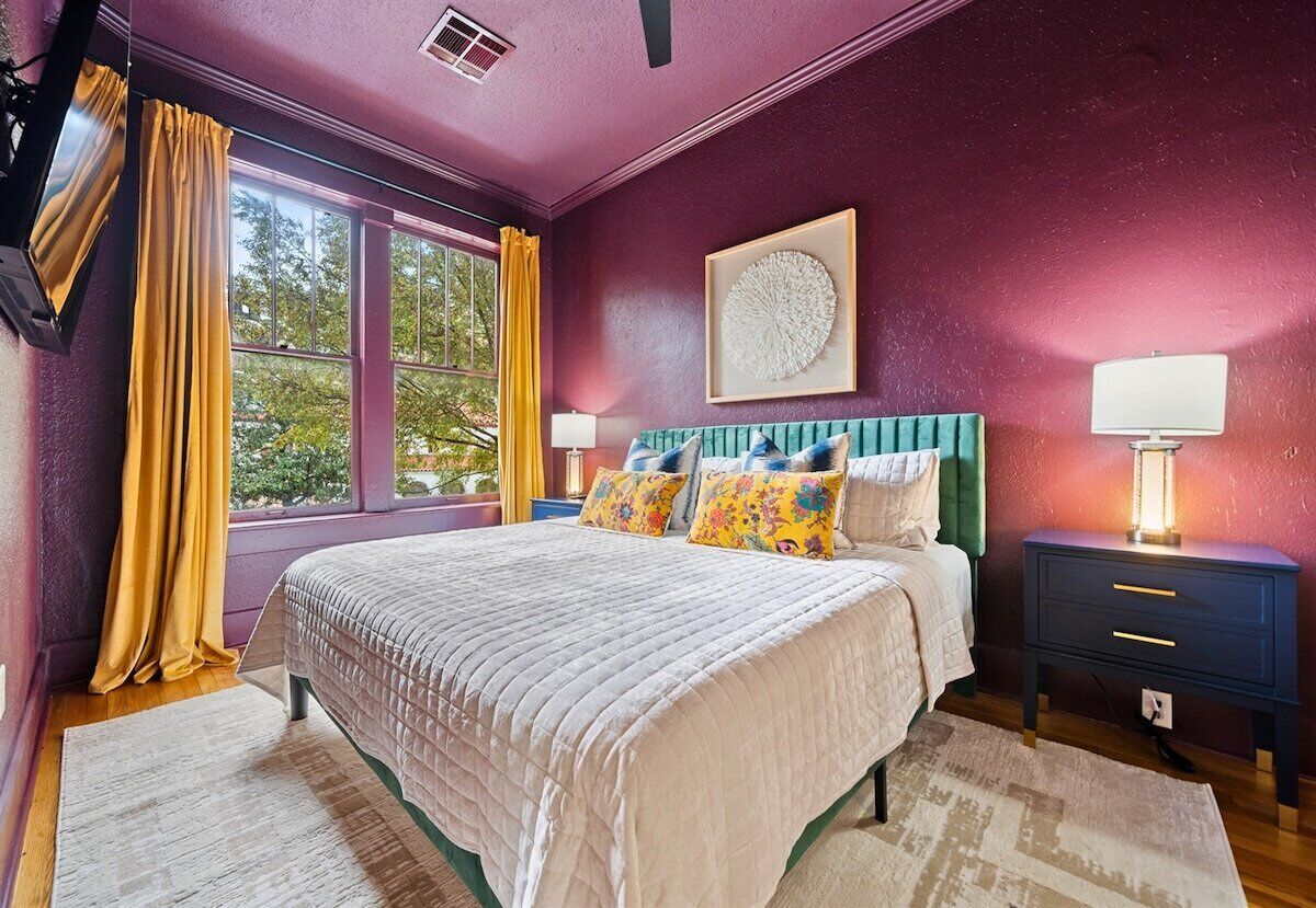 A bedroom with a bed, a nightstand, a lamp, a window with curtains, and a wall-mounted artwork.