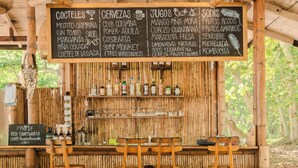 A rustic bar area with a bamboo backdrop, wooden stools, and a menu board listing drinks and snacks. - Santuario Playa Bonita (Santa Marta)