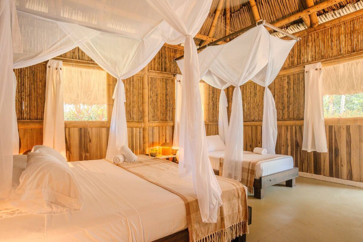 A room with two beds, each with a canopy bedspread and a bedside table with a lamp. The room has wooden walls and a thatched roof.