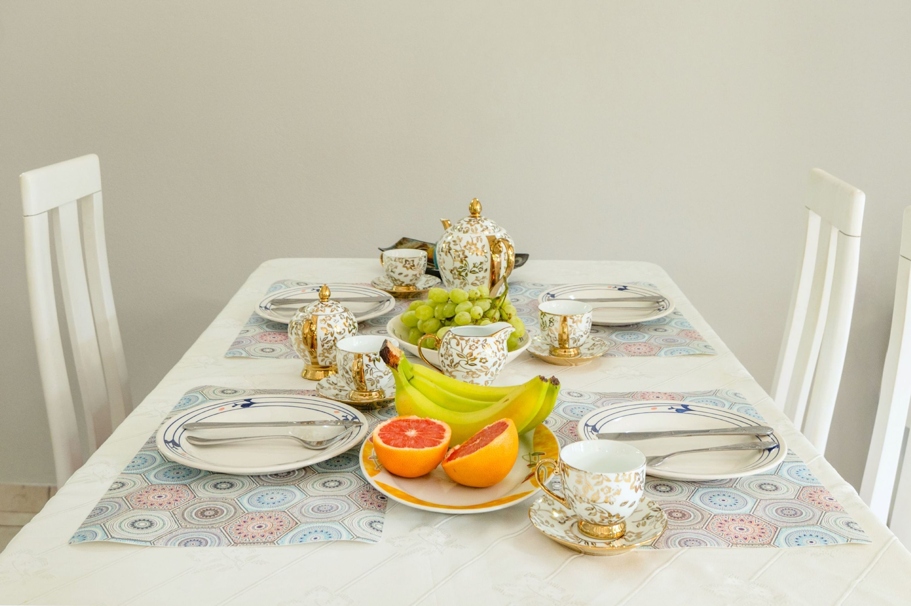 A dining table set with white and gold tableware, a fruit bowl with grapes and bananas, and a half-cut grapefruit.