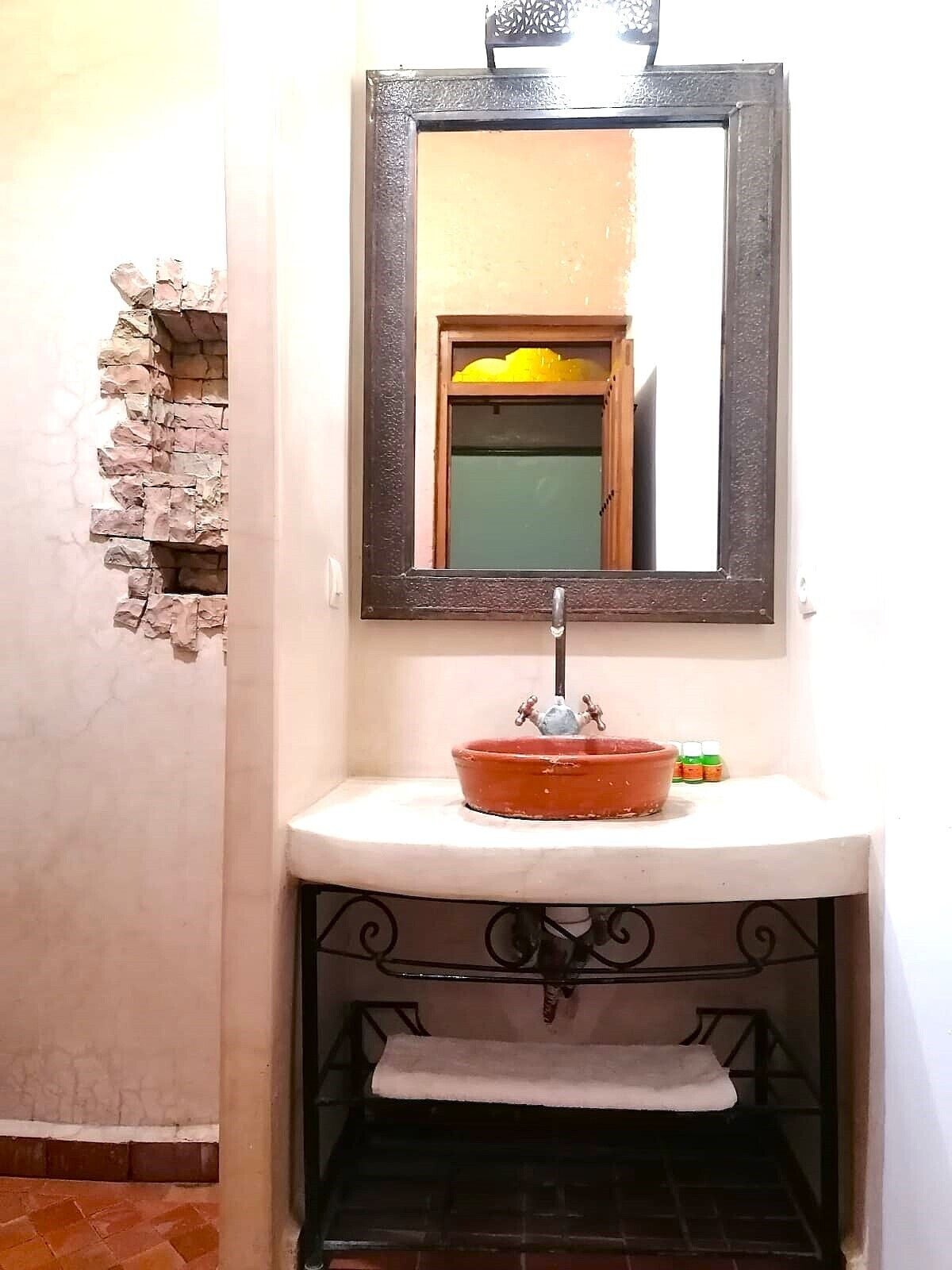 Casa de banho com parede de pedra, espelho, lavatório com cuba vermelha e pequena estante com produtos de higiene pessoal.