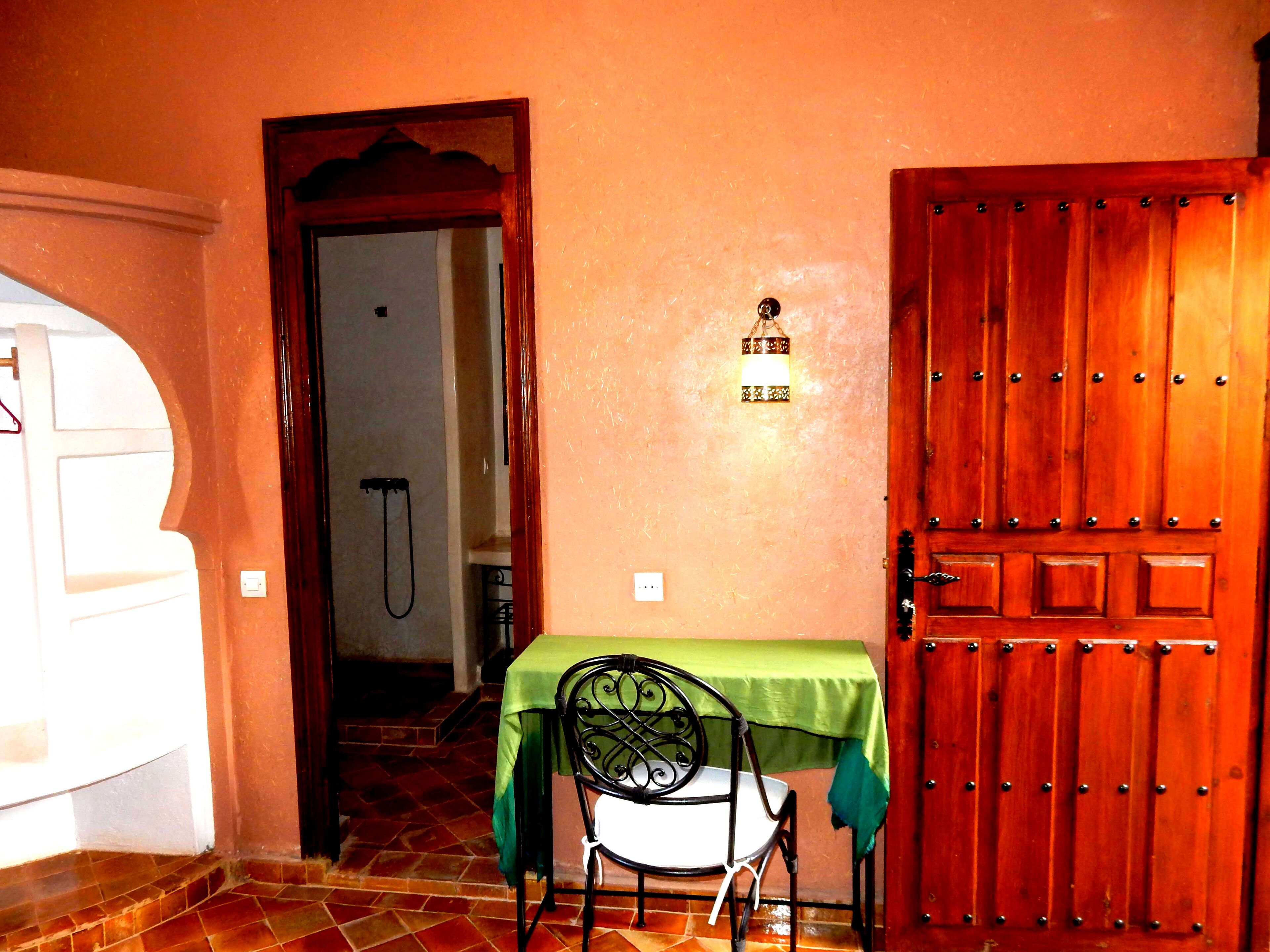Une pièce avec une porte en bois, une table recouverte d’une nappe verte, une chaise et un luminaire fixé au mur.