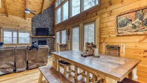Interior acogedor de una cabaña de troncos con chimenea de piedra, muebles de madera y un ventanal con persianas.