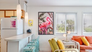A modern kitchen with a white refrigerator, a bar area with teal stools, a green chair, and a red sofa. - Retro-Themed Condo | 2BR | Perfect Location (Myrtle Beach)