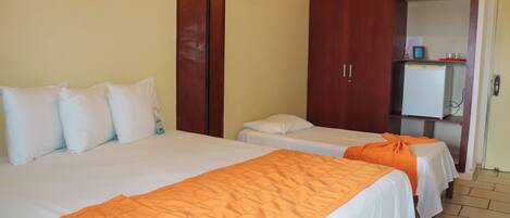 A hotel room with two beds, each with white bedding and orange blankets. There is a wooden wardrobe and a small refrigerator in the corner.
