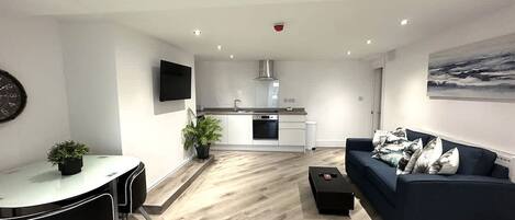 A modern living room with a sofa, a dining table with chairs, a kitchen area with a stove and sink, and a flat-screen TV mounted on the wall.