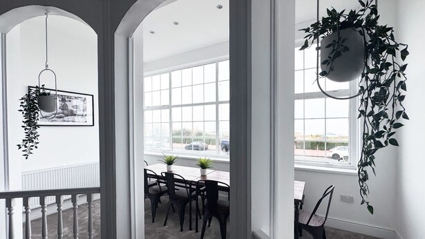 Una habitación luminosa con ventanas amplias, un comedor con mesa y sillas, y plantas colgantes.