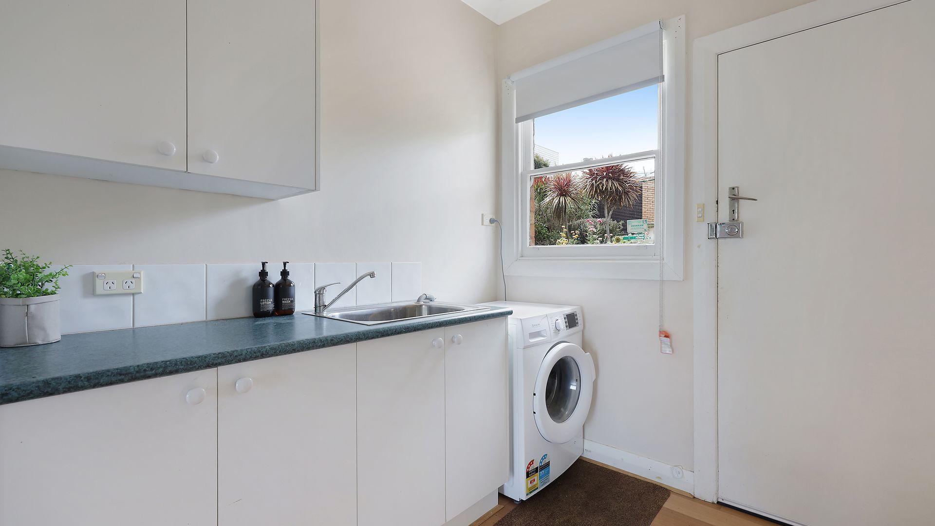 Une cuisine compacte équipée d’un lave-linge, d’un évier et d’une fenêtre donnant sur un espace verdoyant.