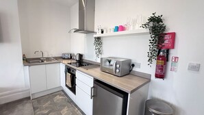 Eine moderne Küche mit weißen Schränken, einer Edelstahl-Dunstabzugshaube, einem Einbauofen, einem Toaster und einem Spülbecken. An der Wand befinden sich Regale mit Gläsern und ein Feuerlöscher.