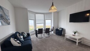Ein modernes Wohnzimmer mit einem dunkelblauen Sofa, einem Esstisch mit Stühlen, einem an der Wand befestigten Fernseher und einem großen Fenster mit Jalousien.