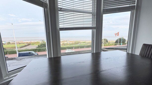 Ein Zimmer mit großem Fenster und Blick auf die Küste, einem dunklen Holztisch und einem einzelnen Stuhl.