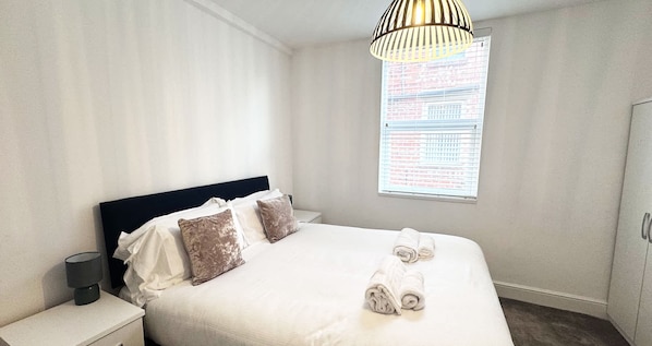 Une chambre d’hôtel comprenant un lit, des oreillers, une table de chevet, une lampe, une fenêtre avec des stores et un luminaire suspendu.