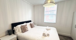 Ein Hotelzimmer mit Bett, Kissen, Nachttisch, Lampe, einem Fenster mit Jalousien und einer Pendelleuchte.
