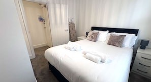 A hotel room with a bed, pillows, a nightstand, a lamp, a door with a sign, and a curtain.